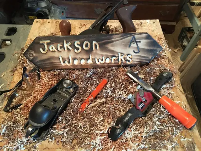 Carved and burned Jackson Woodworks sign on a workbench with hand planes, a chisel and a carpenter's pencil in wood shavings