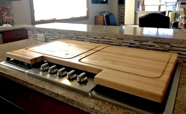 Two fitted hardwood cutting boards covering a stainless gas cooktop on a granite island with a mosaic tile backsplash